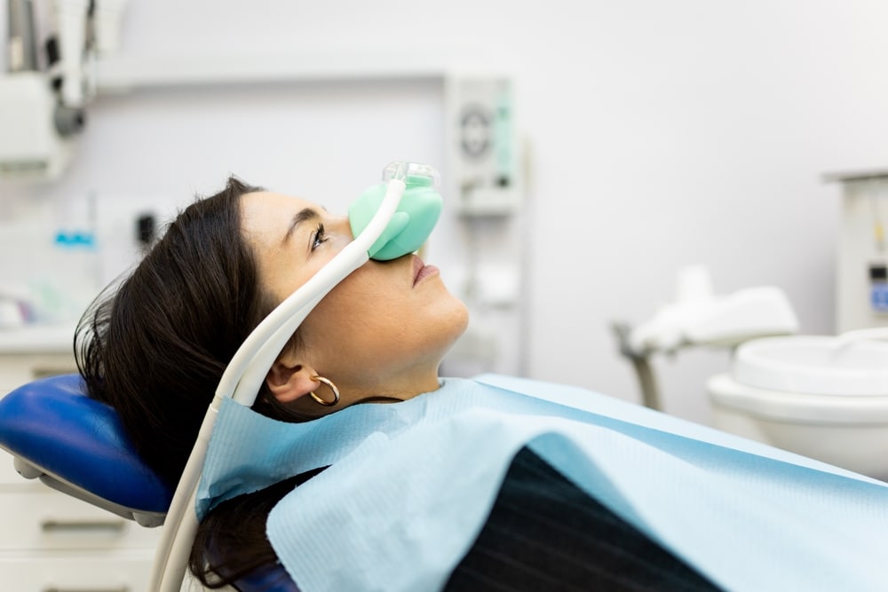 A woman lies in a dental chair with a nasal mask, looking up. Sedation Dentistry at All Valley Dental