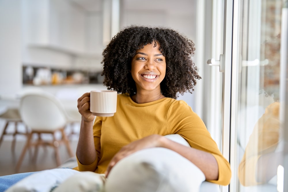 A smiling woman with curly hair holds a mug, looking out a window. Nitrous Oxide at All Valley Dental